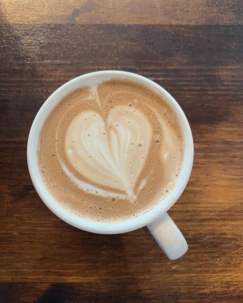 A white mug filled with coffee, topped with frothy milk art shaped like a heart, on a wooden table.