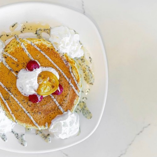A stack of pancakes topped with whipped cream, raspberries, and a slice of lemon, drizzled with syrup on a white plate.