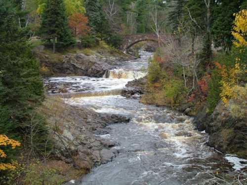 A scenic river flows through a rocky landscape, with a stone bridge and colorful autumn foliage in the background.