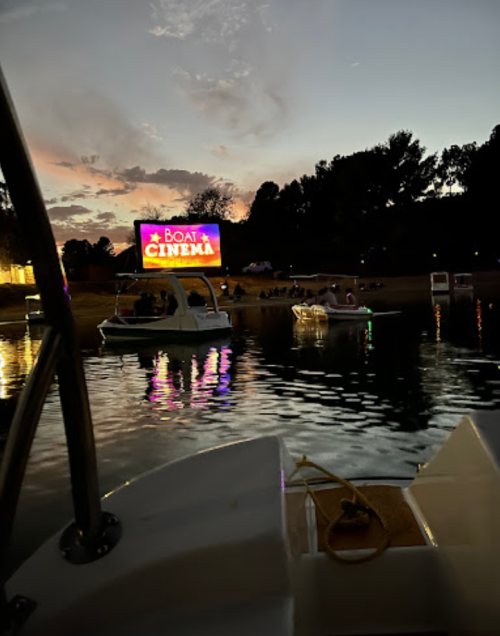 A view from a boat at sunset, featuring a large screen displaying "Boat Cinema" and people enjoying the water.