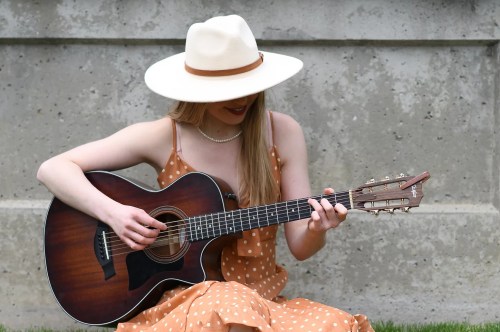 A woman in a polka dot dress and wide-brimmed hat plays an acoustic guitar outdoors.
