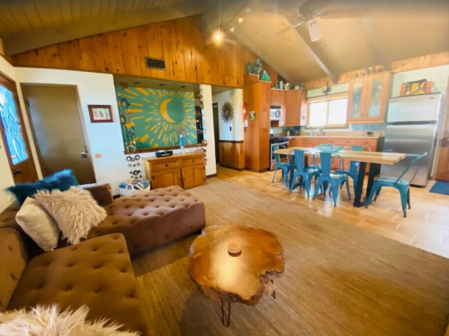 Cozy living area with a brown sofa, wooden table, and a kitchen featuring teal chairs and sunburst decor.