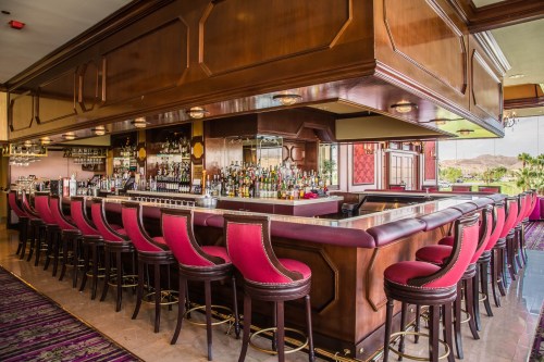 A stylish bar with a polished wooden counter and red upholstered stools, surrounded by shelves of various liquor bottles.