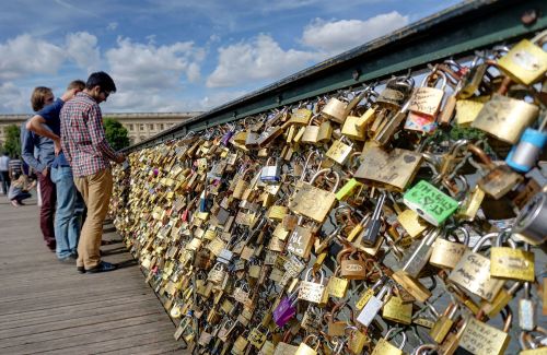 People stand by a bridge covered in colorful love locks, with a blue sky and clouds in the background.