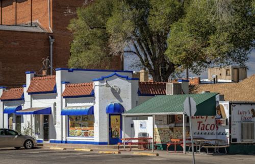 Colorful taco restaurant with blue and white exterior, outdoor seating, and nearby trees in a small town setting.