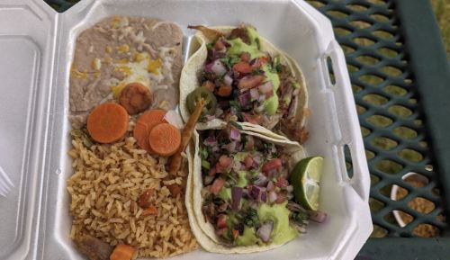 A takeout container with two tacos topped with green sauce, diced tomatoes, rice, refried beans, and sliced carrots.