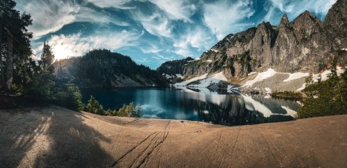 A serene mountain lake surrounded by rocky cliffs and lush trees, reflecting a vibrant sky with wispy clouds.