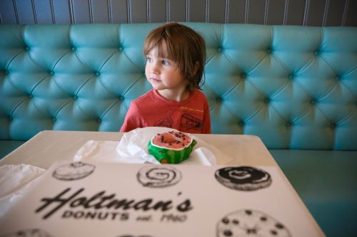 A young child sits at a table with a donut box, looking curiously to the side in a bright, colorful setting.
