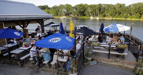 Outdoor dining area by a river, with people seated under umbrellas, enjoying food and drinks on a sunny day.