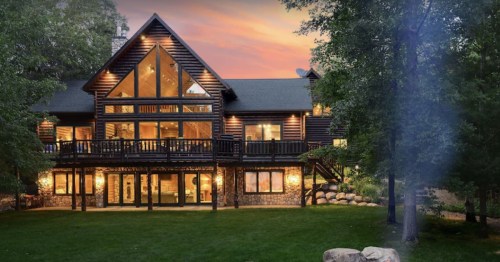 A large log cabin with a stone foundation, surrounded by trees, illuminated at sunset.