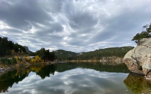 A serene lake surrounded by trees and mountains, reflecting a cloudy sky.