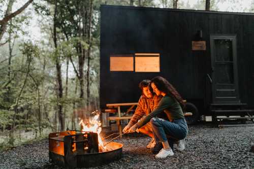 A couple sits by a campfire, roasting marshmallows, with a tiny house in the background surrounded by trees.