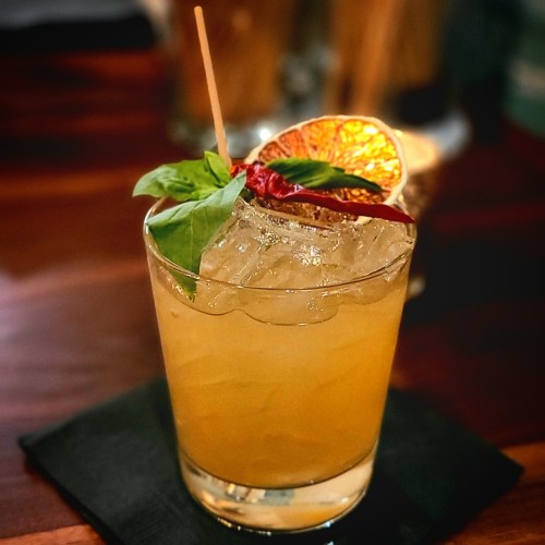 A refreshing cocktail with ice, garnished with a dried orange slice and basil, served on a black napkin.