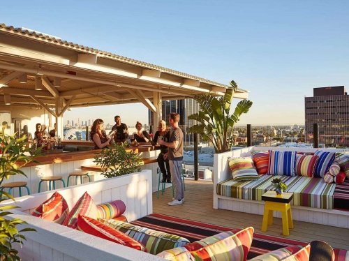 A vibrant rooftop bar with colorful seating, people socializing, and a city skyline in the background during sunset.