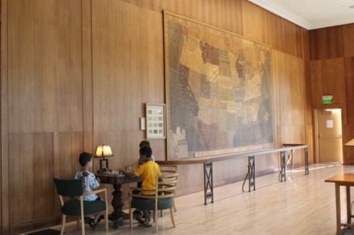 Two children sit at a table in a wooden-paneled room, with a large map of the United States on the wall behind them.