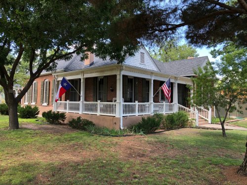 A charming brick house with Texas and American flags, surrounded by greenery and trees.