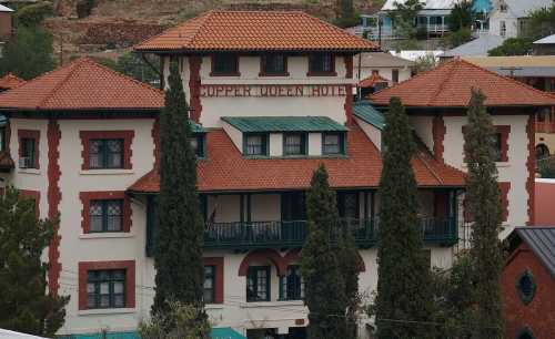 Historic Copper Queen Hotel with red tile roof and tall cypress trees, set against a hillside backdrop.