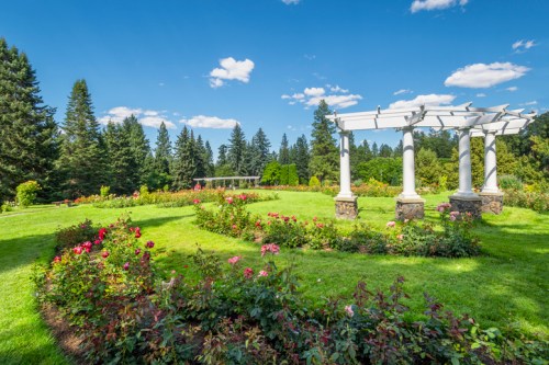 A lush garden with colorful flowers, green lawns, and a white pergola under a blue sky with fluffy clouds.