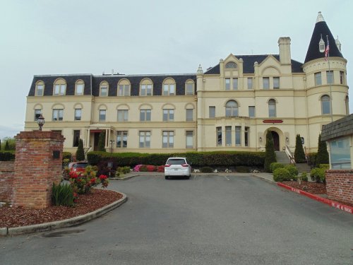 Historic building with a turret, large windows, and landscaped grounds, featuring a parking area in front.
