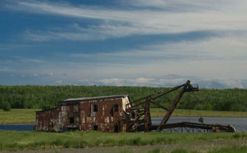 An abandoned, rusted structure partially submerged in water, surrounded by lush green trees and a blue sky.