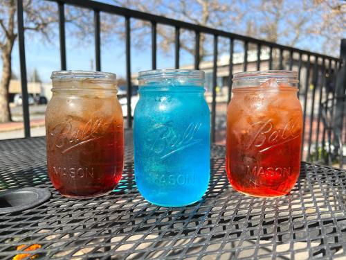 Three mason jars filled with colorful drinks: one brown, one blue, and one orange, sitting on a black table outdoors.