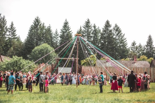 A large group of people dancing around a maypole with colorful ribbons in a grassy field surrounded by trees.