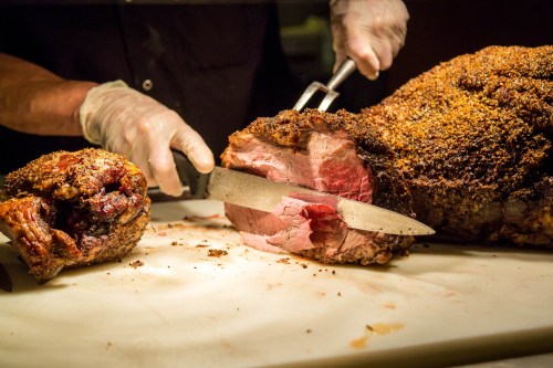 A chef in gloves slices a large, roasted piece of meat on a cutting board, showcasing its juicy interior.