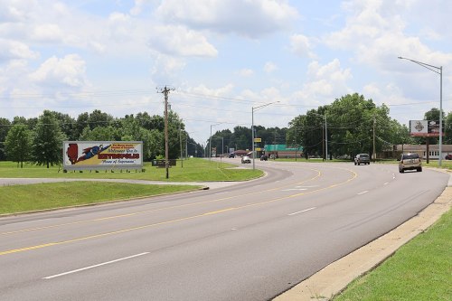 A wide road curves through a green landscape, featuring a billboard and a few cars under a cloudy sky.