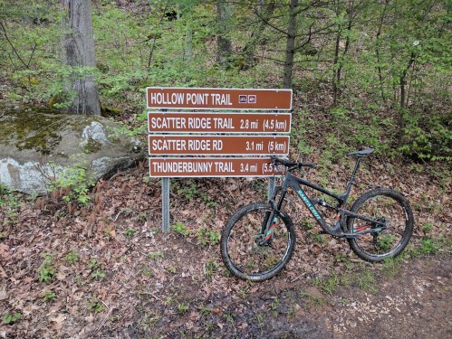 A mountain bike rests beside a trail sign with distances for Hollow Point Trail and other nearby trails in a wooded area.