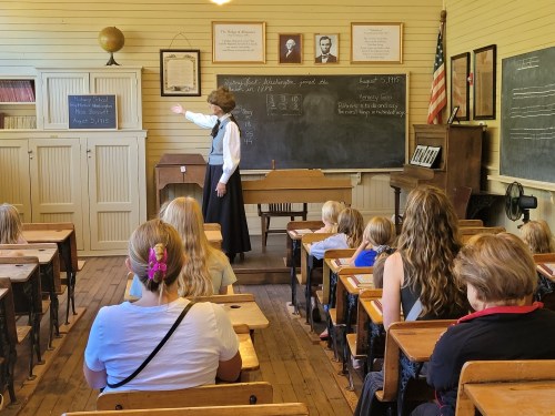 A teacher instructs students in a vintage classroom with wooden desks and chalkboards.