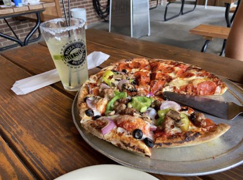 A pizza topped with pepperoni, sausage, olives, and peppers, alongside a drink in a glass on a wooden table.