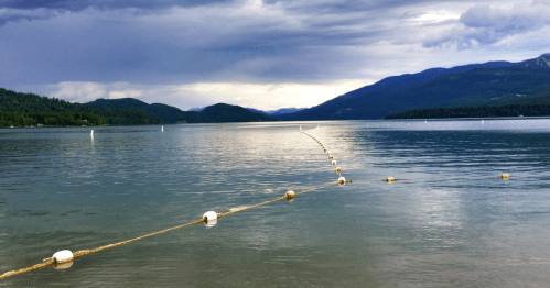 A calm lake with a floating rope barrier, surrounded by mountains under a cloudy sky.