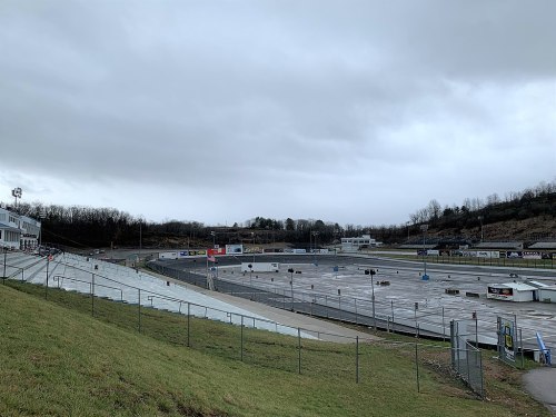 A cloudy day at a racetrack, featuring empty grandstands and a wide, paved track surrounded by green grass.