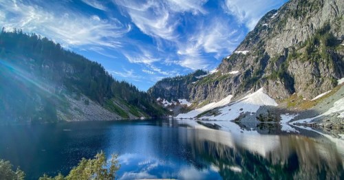 A serene mountain lake surrounded by lush greenery and snow-capped peaks under a blue sky with wispy clouds.