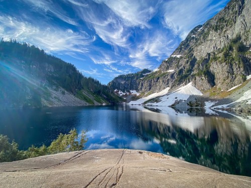 A serene mountain lake surrounded by rocky cliffs and lush greenery, reflecting a blue sky with wispy clouds.