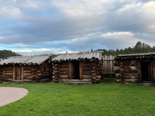 Three rustic log cabins with wooden doors, set against a backdrop of green grass and a cloudy sky.