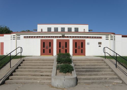 Exterior of a historic swimming pool building with red doors and steps, surrounded by green grass under a clear blue sky.