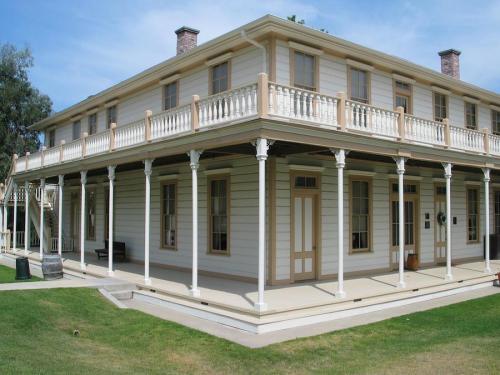 A two-story historic building with a wraparound porch, white siding, and decorative columns set in a grassy area.