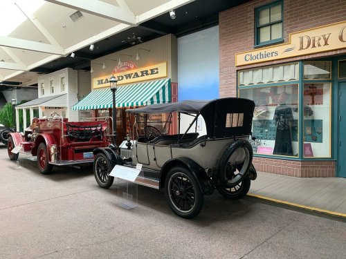 A vintage gray car parked in a museum street, with a red fire truck and storefronts in the background.