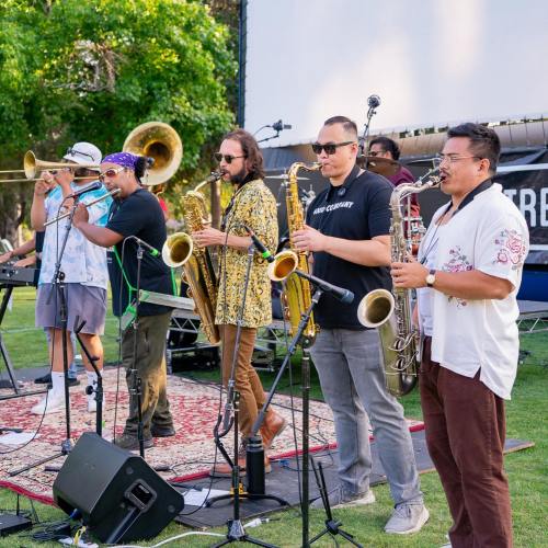 A lively band performs on stage, featuring musicians with brass instruments and a keyboard, set in a sunny outdoor venue.