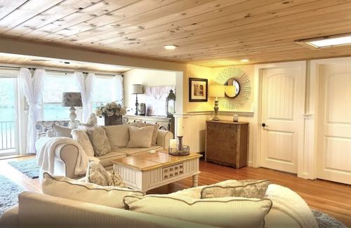 Cozy living room with wooden ceiling, light decor, a comfortable sofa, and large windows letting in natural light.