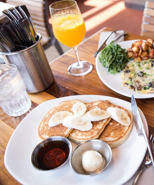 A plate of pancakes topped with banana slices, served with syrup and butter, alongside a mimosa and a vegetable omelet.