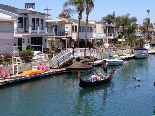 A gondola glides through a calm canal lined with modern homes and palm trees, with boats docked nearby.