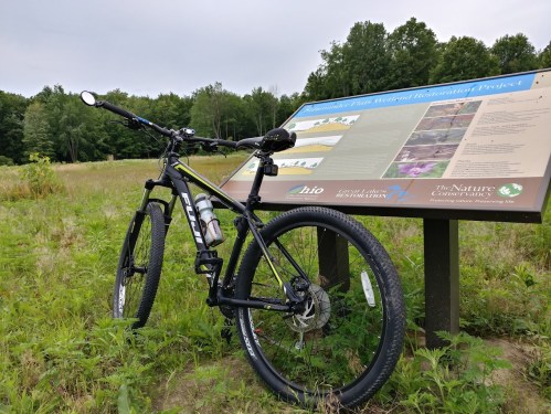 A black mountain bike rests on grass in front of an informational sign about a nature restoration project.