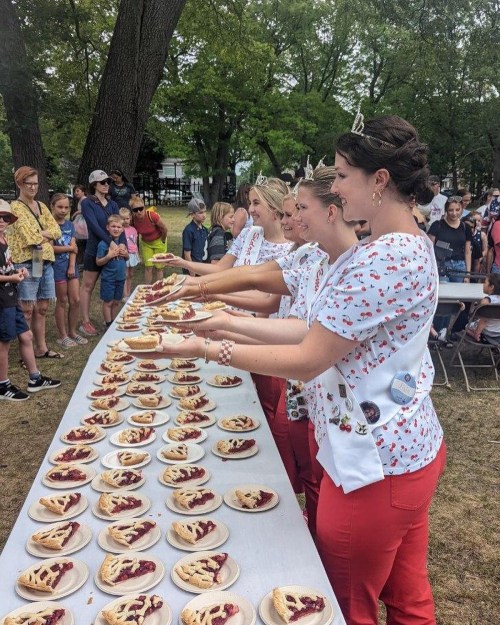 Four women in matching outfits serve pies at an outdoor event, with a crowd of spectators watching nearby.