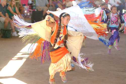 A young girl in colorful traditional attire dances, showcasing vibrant fringed garments at a cultural event.