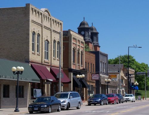 A sunny street scene featuring historic buildings, shops, and parked cars in a small town. A clock tower is visible in the background.