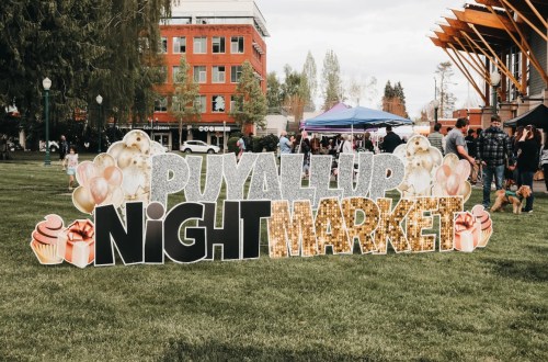 Sign for "Puyallup Night Market" in a park, with people and tents in the background on a cloudy day.
