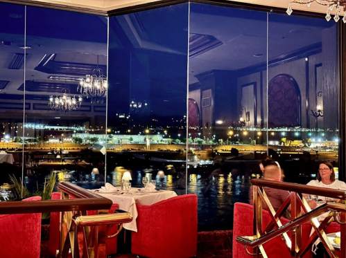 A dimly lit restaurant with red seating, featuring large windows showcasing a city skyline at night.