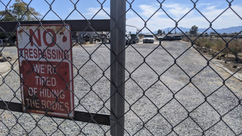 A chain-link fence with a "No Trespassing" sign, warning against entering a deserted area with vehicles in the background.